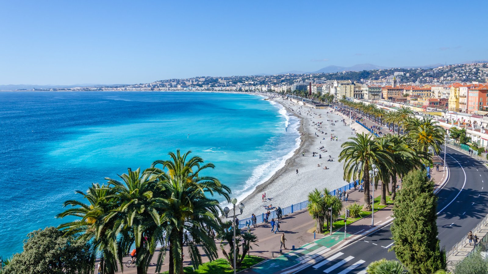 This stunning photograph captures the essence of the French Riviera with Nice's famous Promenade des Anglais framing the turquoise waters of the Mediterranean. Palm trees sway gently in the foreground, evoking a sense of tropical elegance, while the pebbled shore is dotted with beachgoers enjoying the sun-soaked Riviera.