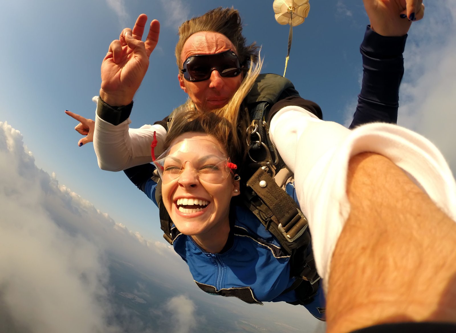 The image captures an adrenaline-fueled moment of sheer exhilaration, with a tandem skydiving duo mid-jump, surrounded by the expansive blue sky. The infectious joy on the woman's face, framed by her windswept hair and goggles, conveys a sense of freedom and abandon that comes with skydiving. The instructor behind ensures a safe and thrilling experience, giving a peace sign as they plummet through the air.