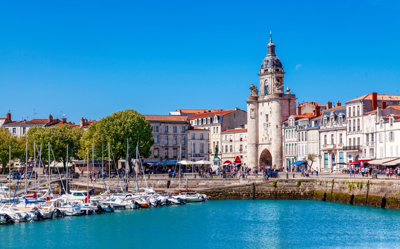 The image captures the historic La Rochelle tower, an enduring symbol of the city's maritime history, overlooking the tranquil waters of the old harbor. Boats moored in the foreground reflect a centuries-old tradition of seafaring, while the bustling promenade echoes the vibrancy of modern life in this storied French port.