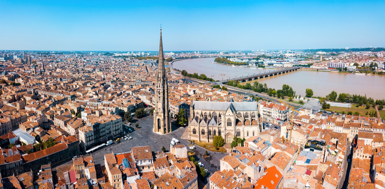 Captured from a high vantage point, this image showcases the towering spire of the Saint-Michel Basilica as it rises above Bordeaux's classical cityscape. The intricate Gothic architecture of the basilica commands the skyline, while the rooftops below unfold towards the Garonne River, which snakes through the city. The clarity of the day offers a pristine view of Bordeaux's urban charm and historical grandeur.