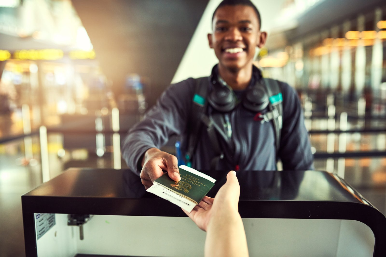 This image presents the quintessential start to any traveler's adventure: the exchange of a passport at an airport check-in counter. In the foreground, a hand extends with a passport, representing the gateway to new experiences. The smiling traveler in the background, equipped with headphones and a backpack, embodies the spirit of youthful anticipation and the eagerness for the experiences that lie ahead.