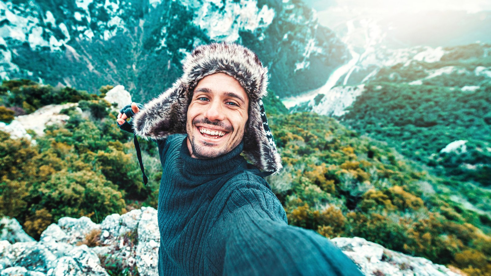 The image showcases an exuberant adventurer taking a selfie at a mountain summit. His joyful expression and the casual, outstretched pose with a selfie stick create a sense of connection with viewers, inviting them to share in the triumph of his ascent. Clad in a cozy sweater and a fur-lined hat against the crisp mountain air, he embodies the spirit of adventure travel. The scenic backdrop offers a stunning blend of lush greenery and rugged terrain, highlighting the beauty of the natural world.