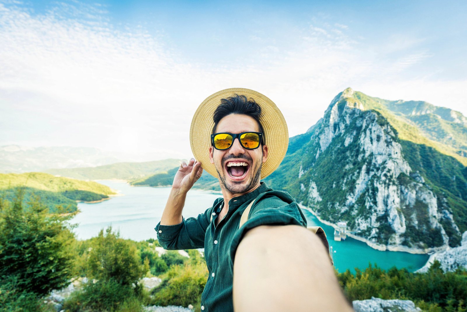 This image is brimming with the exhilaration of adventure as a cheerful man takes a selfie against the backdrop of majestic mountains. His wide grin, stylish sunglasses reflecting the sky, and the casual tip of his straw hat convey a spirit of carefree exploration and the pleasure of reaching new heights. The vivid green hills rolling into the distance and the serene blue waters below celebrate the natural beauty of the great outdoors.