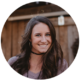 This inviting portrait captures a woman with a radiant smile and sparkling eyes that suggest a friendly and welcoming personality. Her long, dark hair frames her face beautifully, and the subtle glow of the warm lighting complements her casual yet stylish outfit, complete with a chic choker. The natural backdrop adds a relaxed and rustic charm to the image, highlighting her modern elegance.