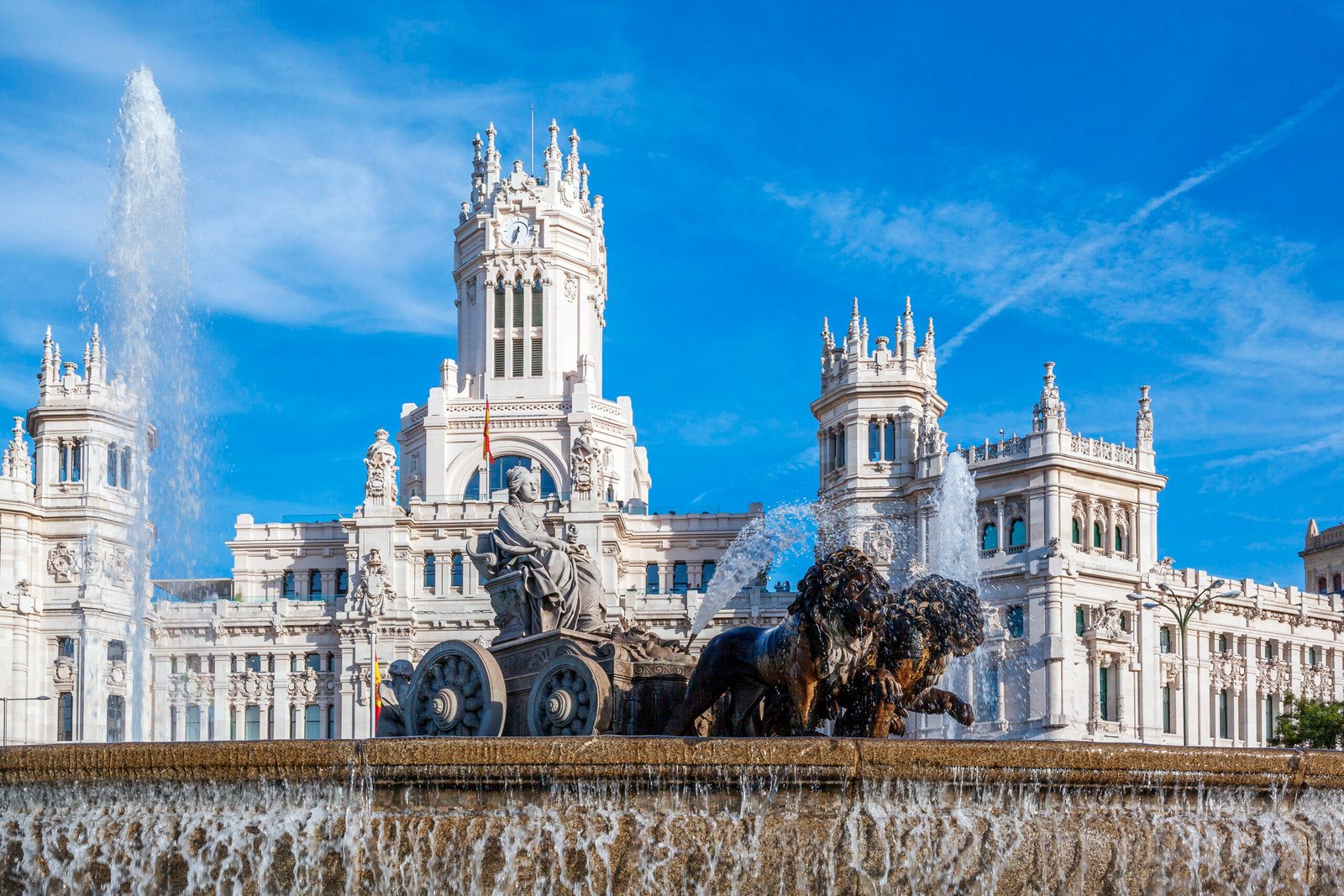 This image captures the grandeur of the Cibeles Fountain, a symbol of Madrid's rich history and artistry. The fountain's detailed sculpture of the goddess Cybele on a chariot pulled by lions stands as a tribute to the city's cultural heritage. In the background, the Palacio de Cibeles's intricate architecture provides a stunning example of early 20th-century design, its white façade gleaming under the clear blue sky.